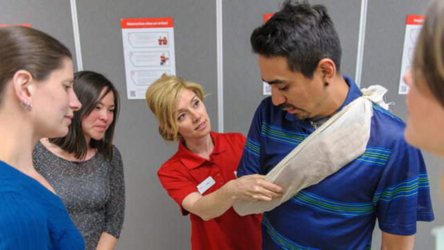 Person wearing a Red Cross Instructor shirt standing in a classroom speaking to two learners.