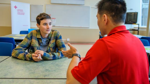 Un moniteur de secourisme discute avec une personne apprenante assise devant lui dans une salle de classe.