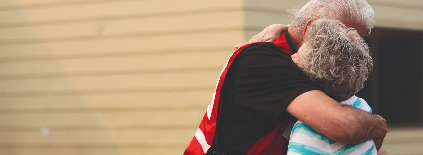 An elderly red cross volunteer provides a hug to an elderly person.