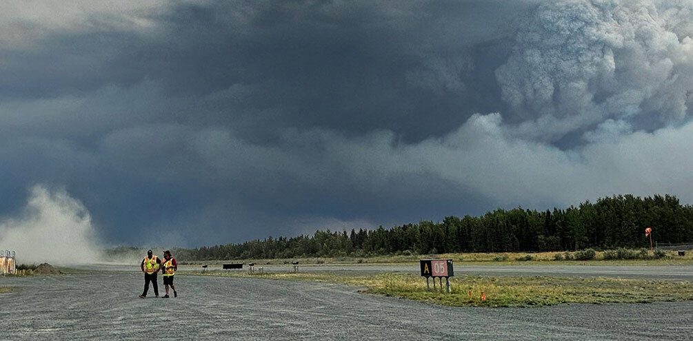 Two people in high-vis vests walk across an airfield as smoke rises from a forest behind them.