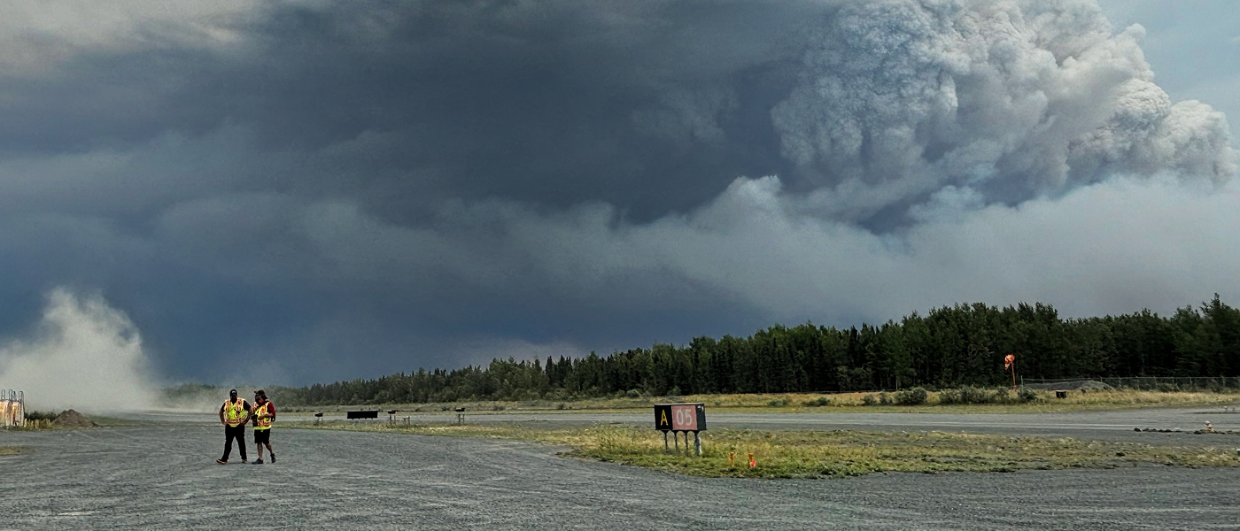 Two people in high-vis vests walk across an airfield as smoke rises from a forest behind them.