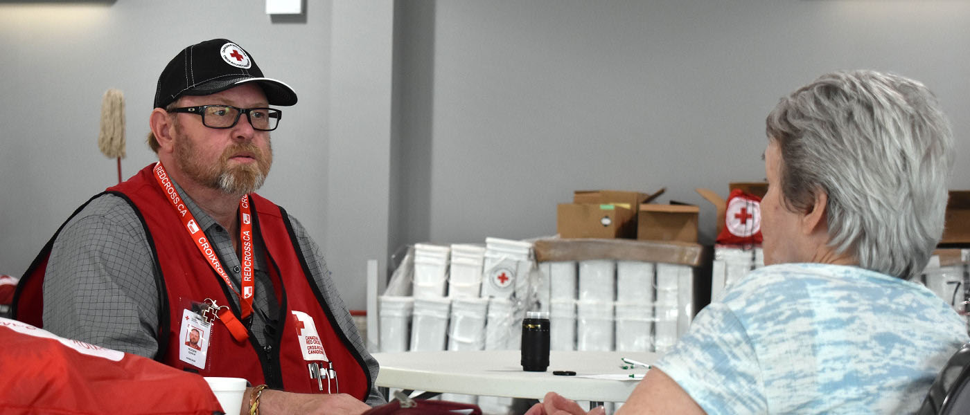 Un homme portant un gilet et une casquette de la Croix-Rouge canadienne écoute une femme aux cheveux gris, qui est dos à la caméra. Ils sont assis à l’intérieur, à une table grise.