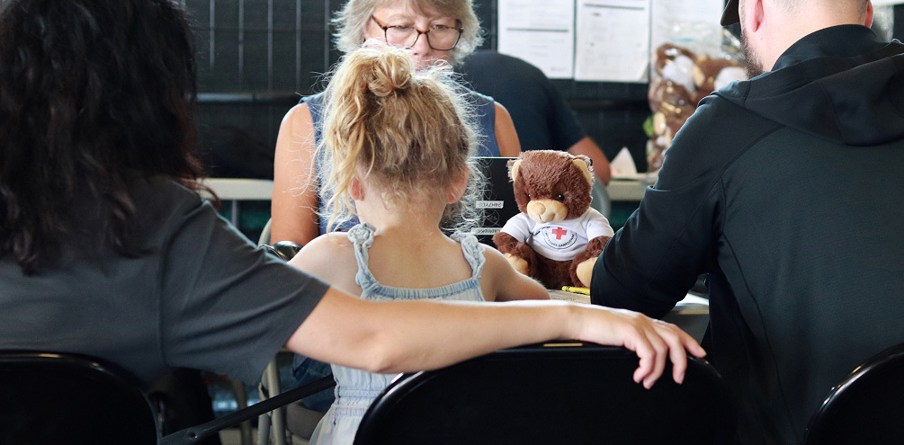 A little girl sits between her parents at a table with a Red Cross teddy bear, while a volunteer provides assistance.