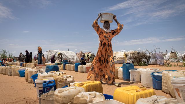 A woman carries a container on her head, surrounded by water containers.