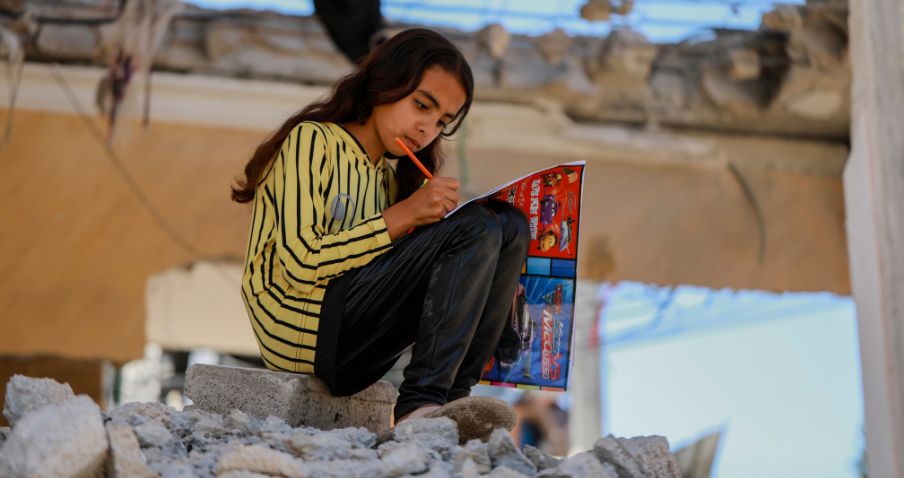 Un enfant assis sur un tas de débris rocheux, écrivant dans un livre, entouré de gravats.