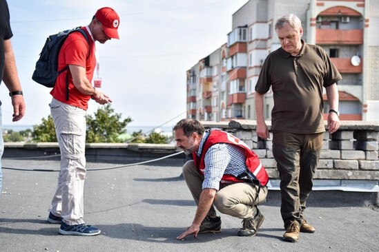 Un homme portant un dossard rouge est accroupi pour toucher le sol alors que deux hommes debout de chaque côté le regardent.