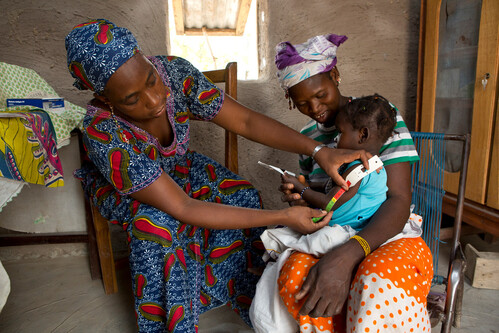 Woman holds a baby as another woman measures the baby’s arm