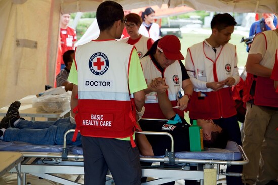 A team of medical delegates wearing Red Cross vests treating a patient on a bed