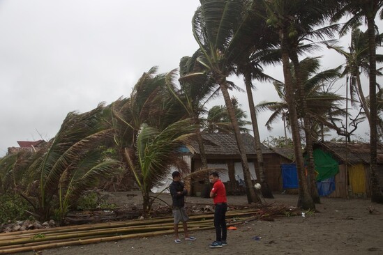 Two people stand in front of a house with strong winds blowing palm trees