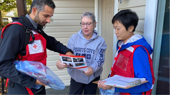 Two people wearing red cross vest show an older adult a prepardness resource at her front door.