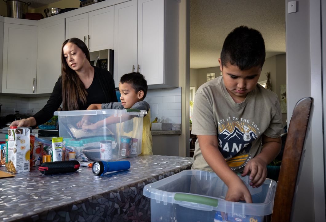 A mother and two children pack an emergency kit. There are two plastic containers full of emergency preparedness goods.