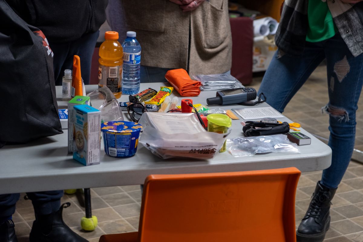 The contents of an emergency kit are laid out on a table including drinks, candy bars, walkie-talkies, matches, emergency blankets, and more. 