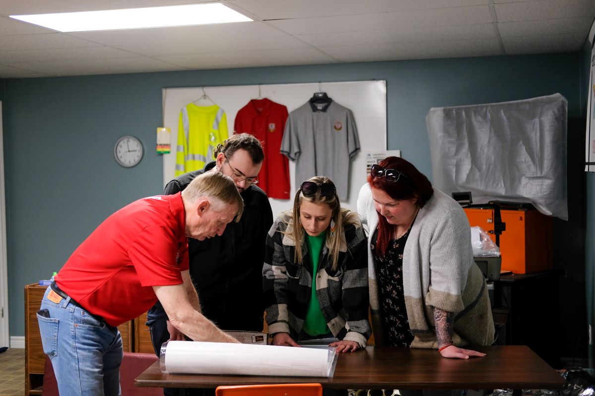 Four people stand looking over a map on a table. One man is pointing to something on the map.