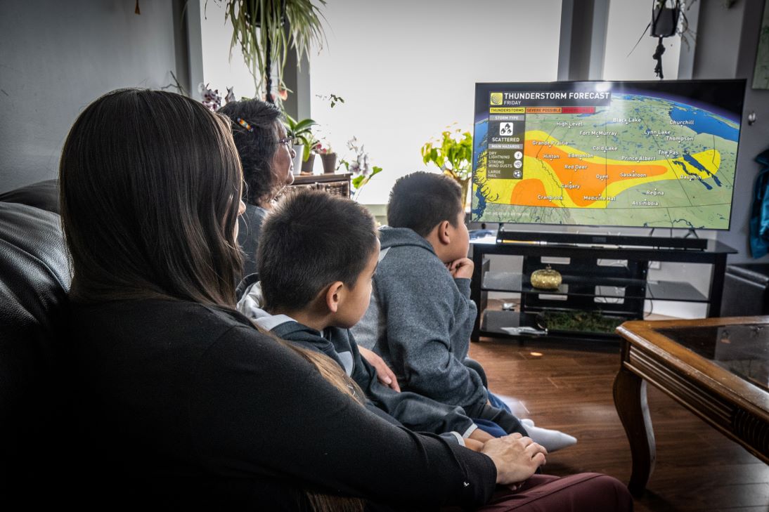 Une grand-mère, une mère et ses deux fils regardent un bulletin météorologique à la télévision. On peut voir à l’écran une carte de différentes couleurs.