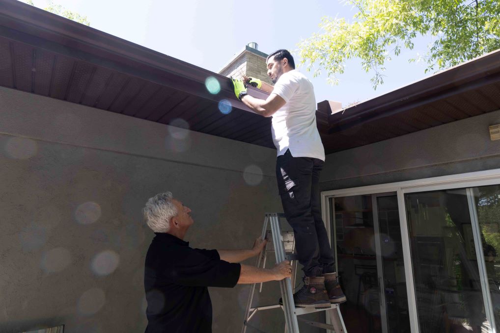 An older man holds the ladder for a man who is cleaning the gutters to remove debris. (Credit John Falcon)