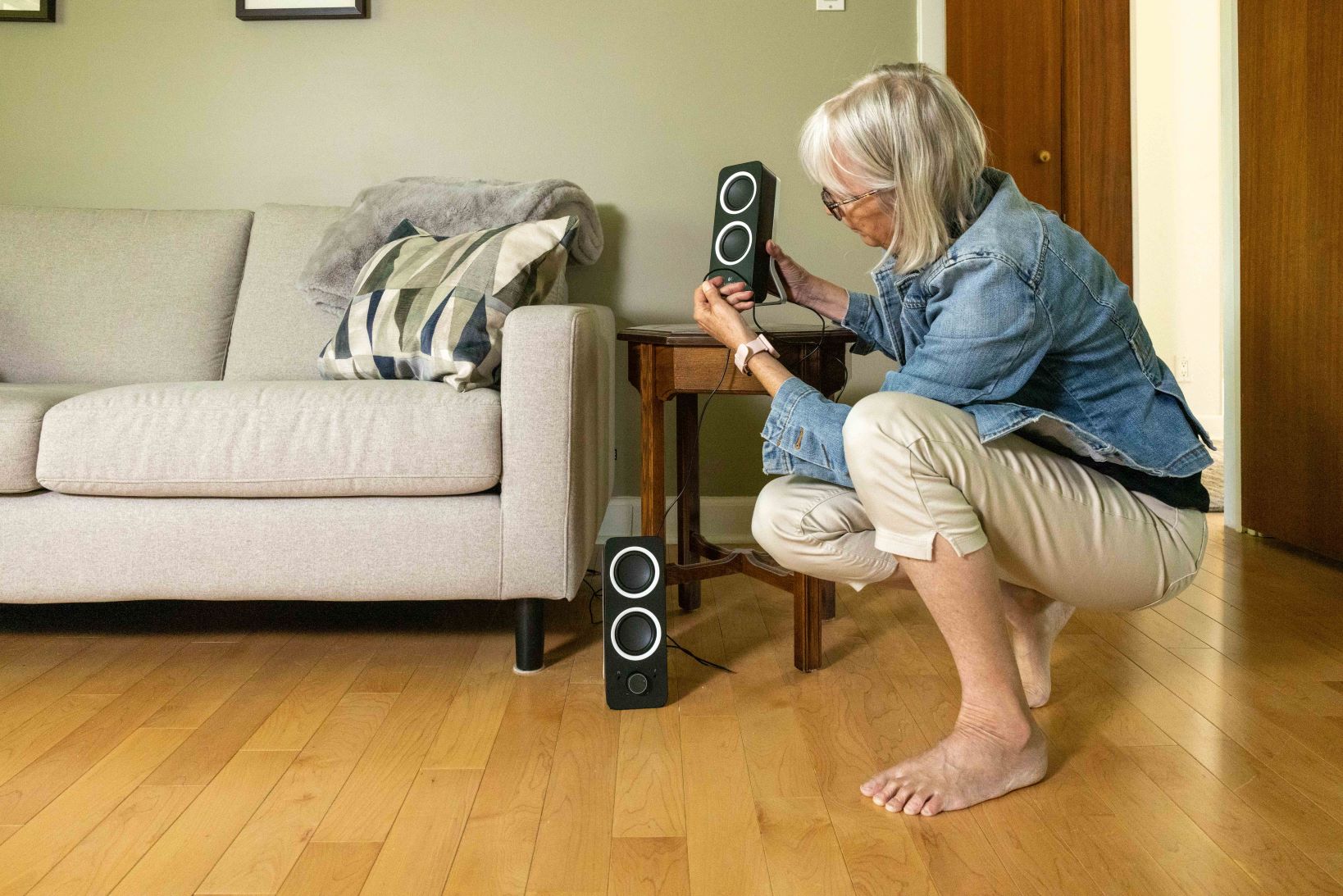 An older woman picks up electronic speakers off the living room floor and places them on a side table. (Credit John Falcon)