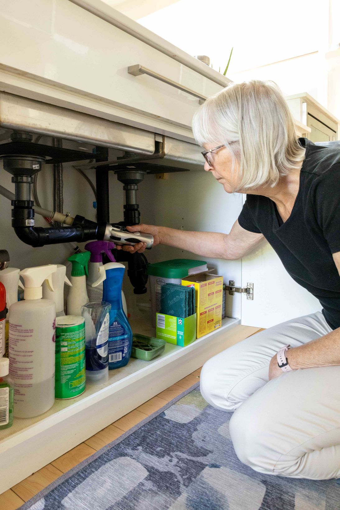 An older woman looks at the pipes under the sink with a flashlight to ensure there are no leaks. (Credit John Falcon)