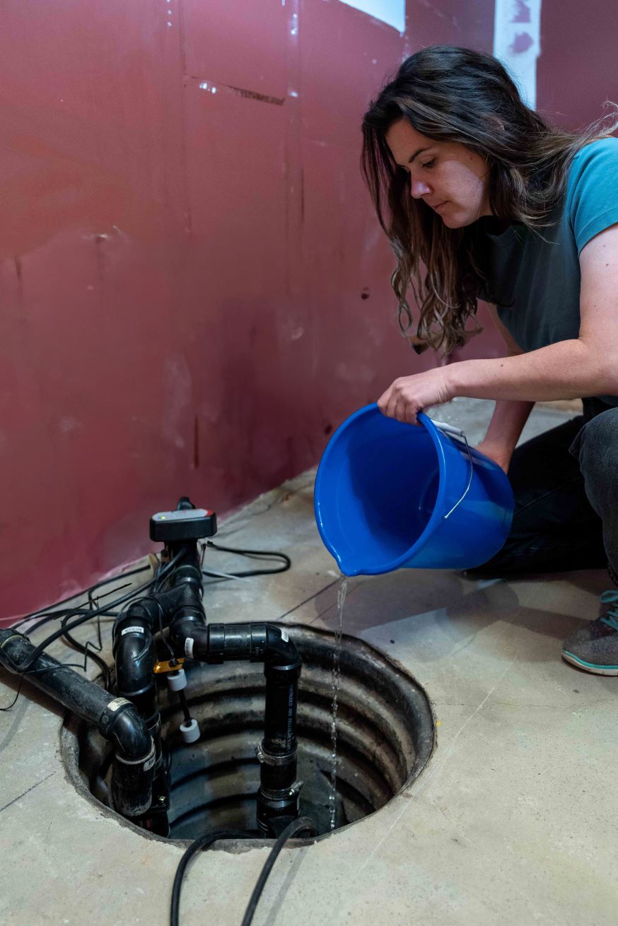 A woman pours water from a bucket into a sump pump. (Credit John Falcon)