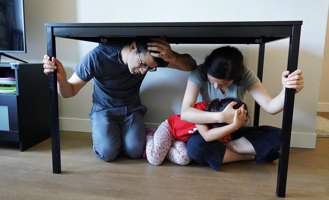 A man, woman, and child take cover under a table during an earthquake. The man and woman are holding onto the table with one hand.