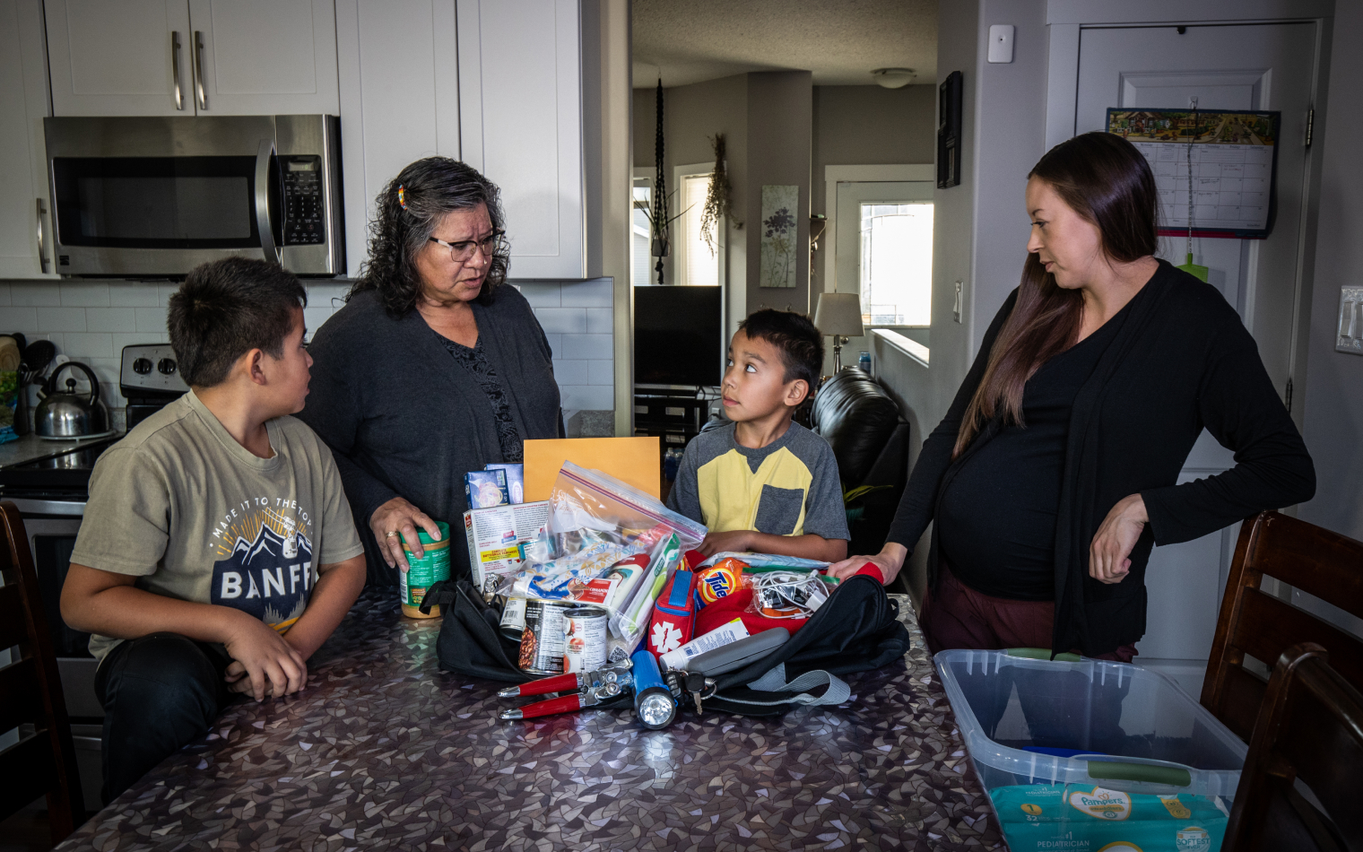 A grandmother, mother and two children stand around a table preparing an emergency kit, items displayed on the table.