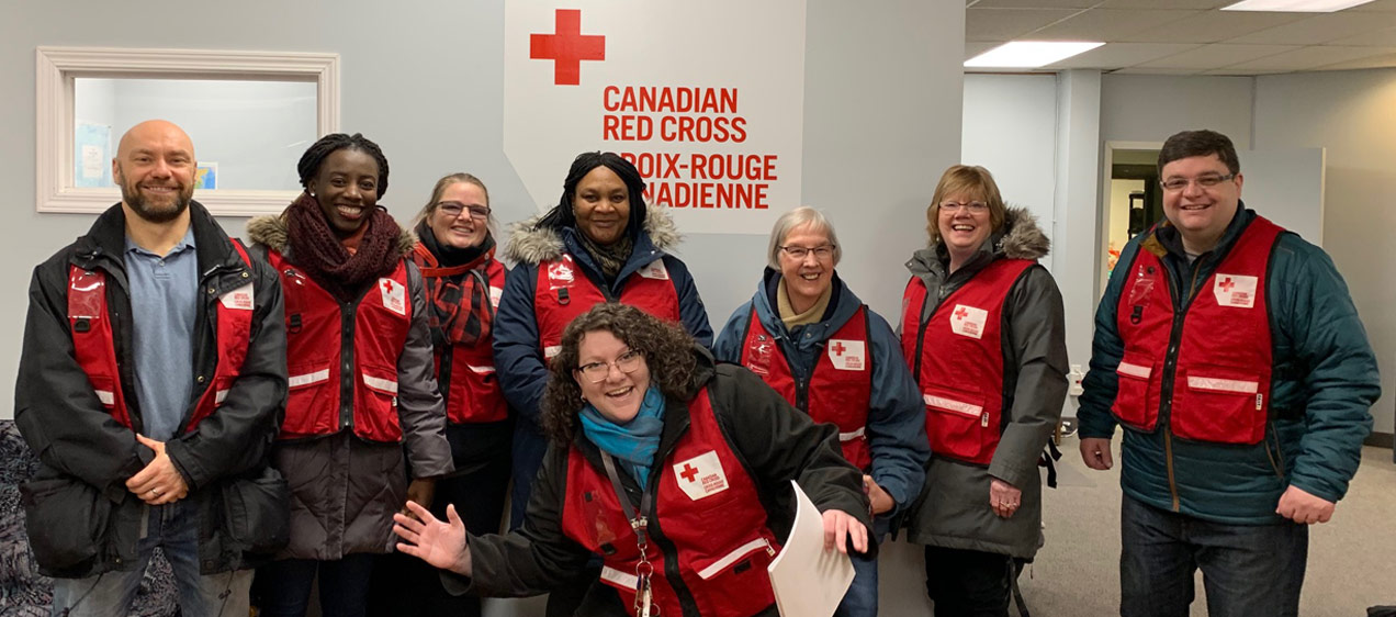 Eight Red Cross volunteers wearing red vest smile as they prepare to help those in need, inside of a building.