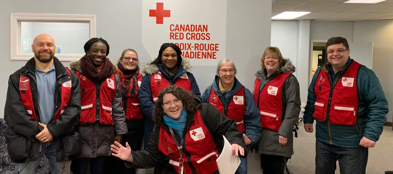 Eight Red Cross volunteers wearing red vest smile as they prepare to help those in need, inside of a building.