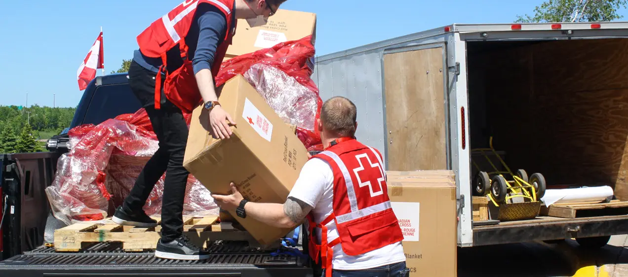 Two male Red Cross volunteers wearing red vests unload emergency supplies from a large truck.