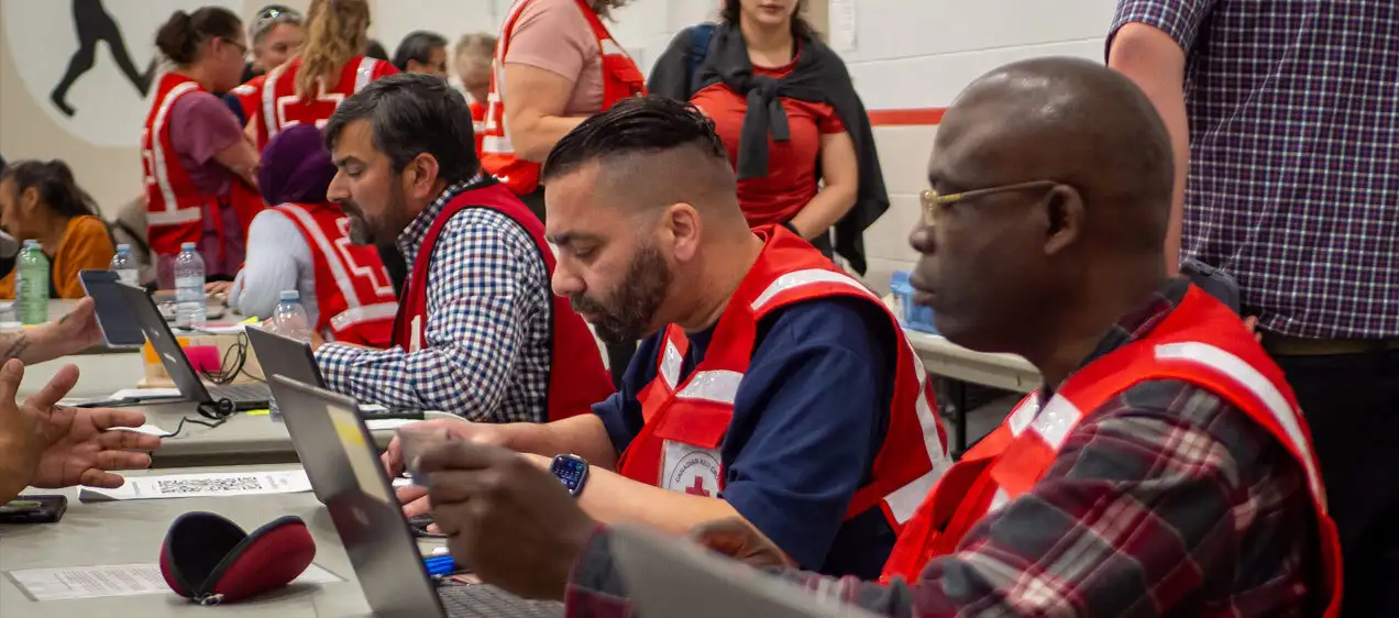 Three Red Cross volunteers with laptops open are helping the community affected by the 2025 Saskatchewan wildfires with emergency services like registration, and personal services.