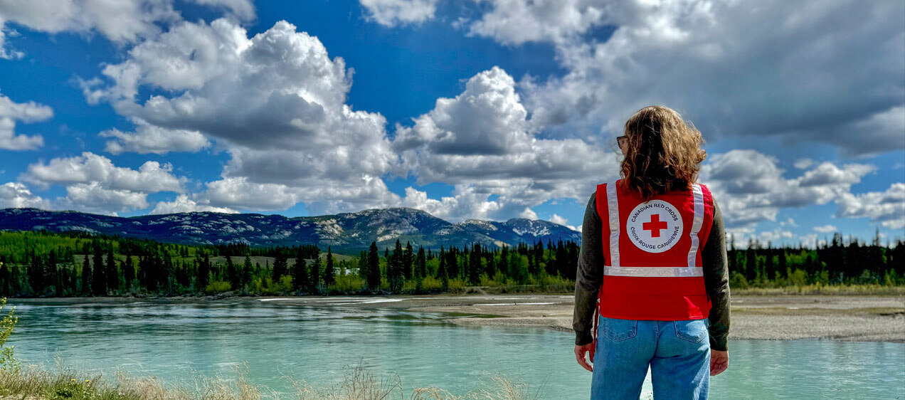 A person in a red Canadian Red Cross vest faces a body of water, with trees and mountains in the distance.