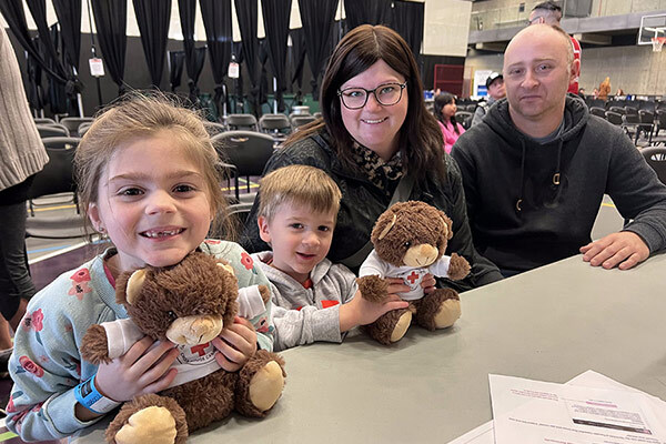 A family of four smiles at the camera. The two children are holding teddy bears.