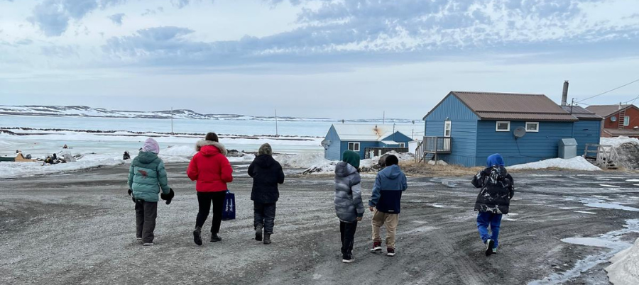 A group of youth walk together on a snowy path in Sanikiluaq, Nunavut, with colorful houses and snow-covered mountains in the background.