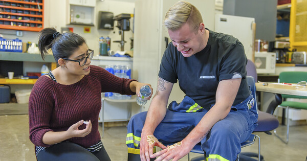 A maintenance worker has a burn on his hands, and a coworker provides first aid on the burn area.