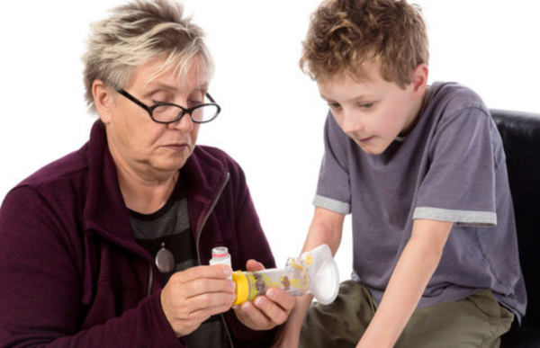 Une adulte aide un enfant asthmatique à utiliser un inhalateur. L’adulte place l’inhalateur dans la chambre de retenue et rassure l’enfant tout au long du processus.