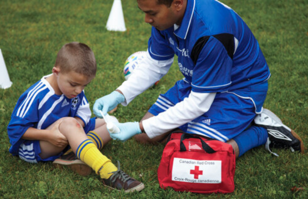 A soccer coach is tending to a child's injured knee by placing a gauze pad on the wound while the boy holds his knee.