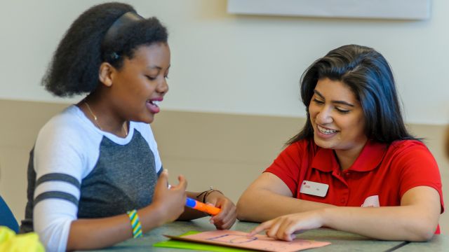 Red Cross personnel engaging with youth, pointing at a drawing while sitting at a table.