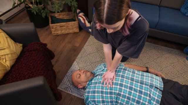A young person kneels next to an older person laying unresponsive on the floor of a living room and performs chest compressions.