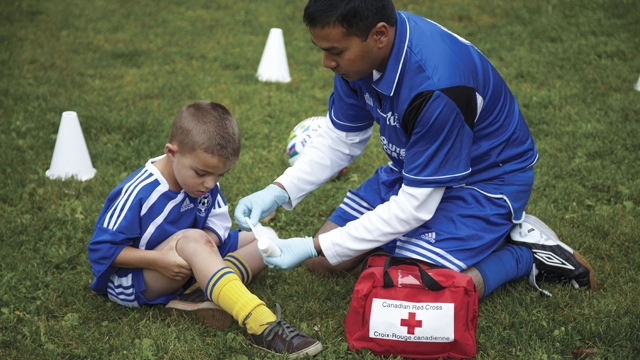 A soccer coach aids a child in soccer gear, treating both knees on the grass with a first aid kit.