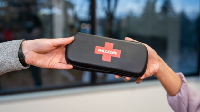 The hands of a person handing a Naloxone kit to another person.