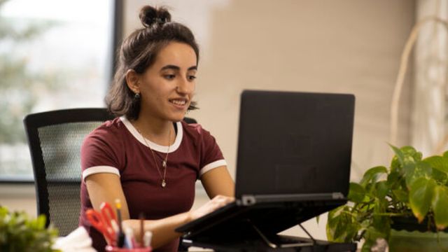 Une personne souriante travaille à un ordinateur portable; le bureau contient une chaise noire, un bureau brun, un porte-crayon et des plantes vertes.