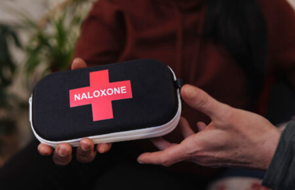 Close-up of a person holding a packaged naloxone nasal spray kit while
gesturing with the other hand, seated at a table across from another person in
a blurred indoor setting.