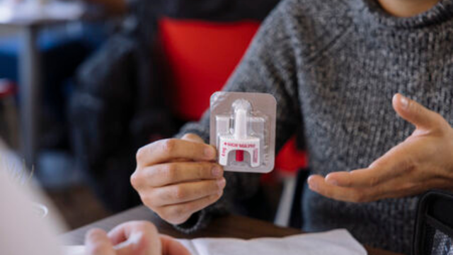 Close-up of a person holding a packaged naloxone nasal spray kit while    gesturing with the other hand, seated at a table across from another person in a blurred indoor setting.