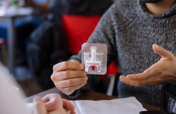 Close-up of a person holding a packaged naloxone nasal spray kit while gesturing with the other hand, seated at a table across from another person in a blurred indoor setting.