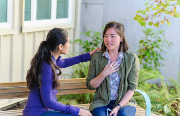 Person sitting on a bench holding their chest and struggling to breathe while another person approaches