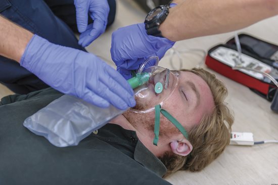 An unresponsive person is being given supplemental oxygen with a non-rebreather mask while someone adjusts the elastic band around the head.