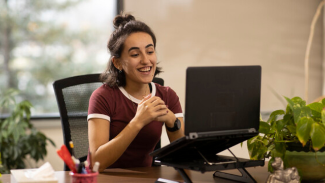 A person working remotely at home smiles at the camera on his desktop computer while clasping hands