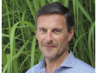  A headshot of a man in a light blue collared shirt, standing in front of a background of tall grass.  
