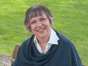  A woman in a dark blue shawl sitting on a bench, open book in hand as she smiles for the camera.  