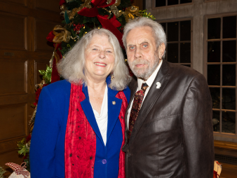  A woman and man pose for a photo, a Christmas tree and large window behind them.  