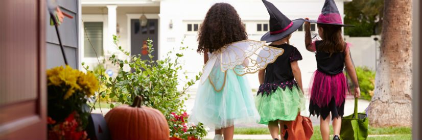 Three children in Halloween costumes are leaving their house to go trick-or-treating, each carrying a bag.