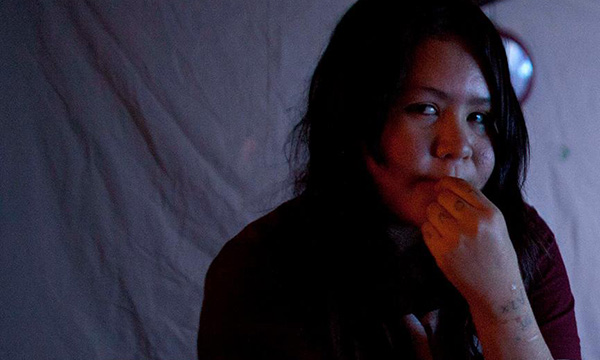 An Indigenous youth holds their hand to their mouth, looking across the room.